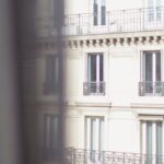 Historic Affordable Housing - Hotel with elegant balconies and bay windows in Paris as seen from opposite building through slightly ajar window curtains on sunny day