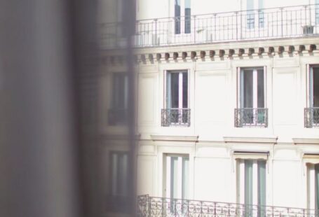 Historic Affordable Housing - Hotel with elegant balconies and bay windows in Paris as seen from opposite building through slightly ajar window curtains on sunny day