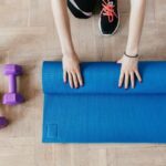 Youth Centers - Top view of anonymous female athlete in black leggings and sneakers unfolding blue mat for exercising on floor near dumbbells and water bottle in modern fitness center