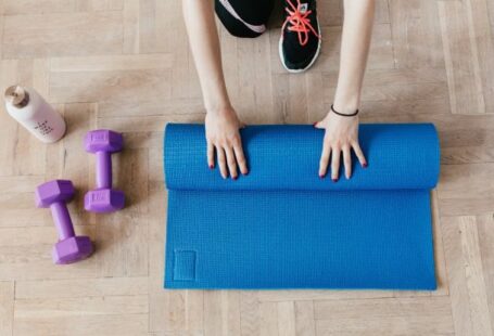 Youth Centers - Top view of anonymous female athlete in black leggings and sneakers unfolding blue mat for exercising on floor near dumbbells and water bottle in modern fitness center